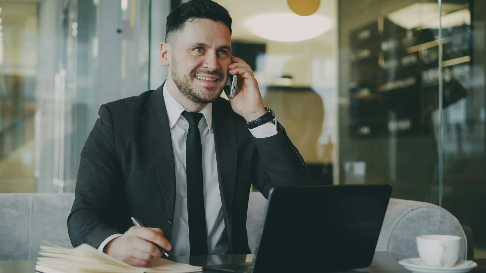 A suited advisor smiling on a phone call while taking notes at a desk.