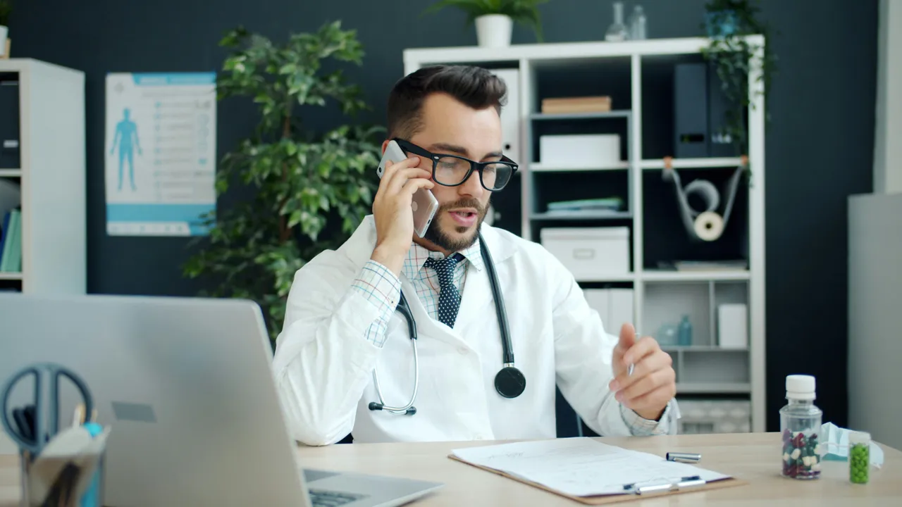 A doctor in a clinic taking a call at their desk, stethoscope around their neck.
