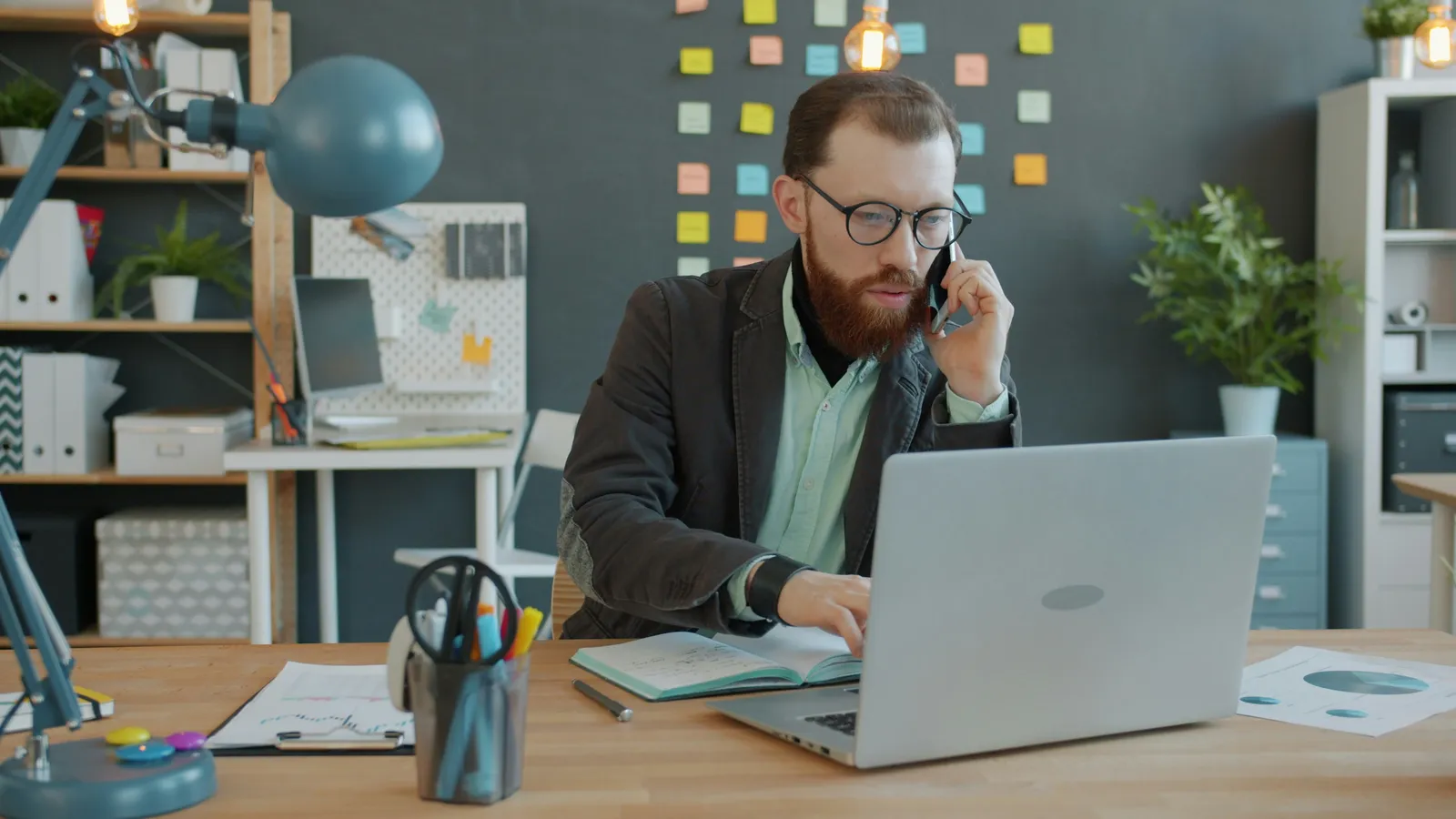 A bearded professional in glasses on a phone call while working at a laptop.